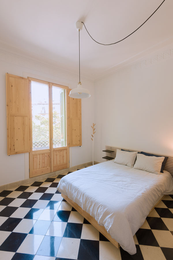 Bright bedroom with checkered floor tiles, wooden shutters opening onto a balcony,a double bed and a vintage white pendant light.