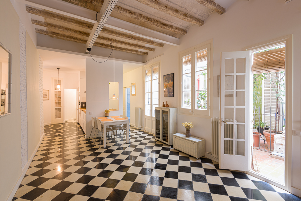 Open-plan dining area with checkered floor tiles, exposed beams, and French doors leading to a patio with potted plants.