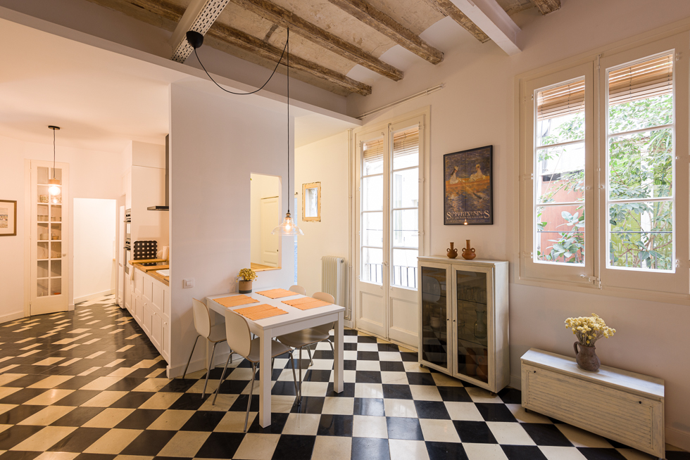 Dining area with checkered floor tiles, exposed beams, a white table with four chairs, and large windows overlooking greenery.