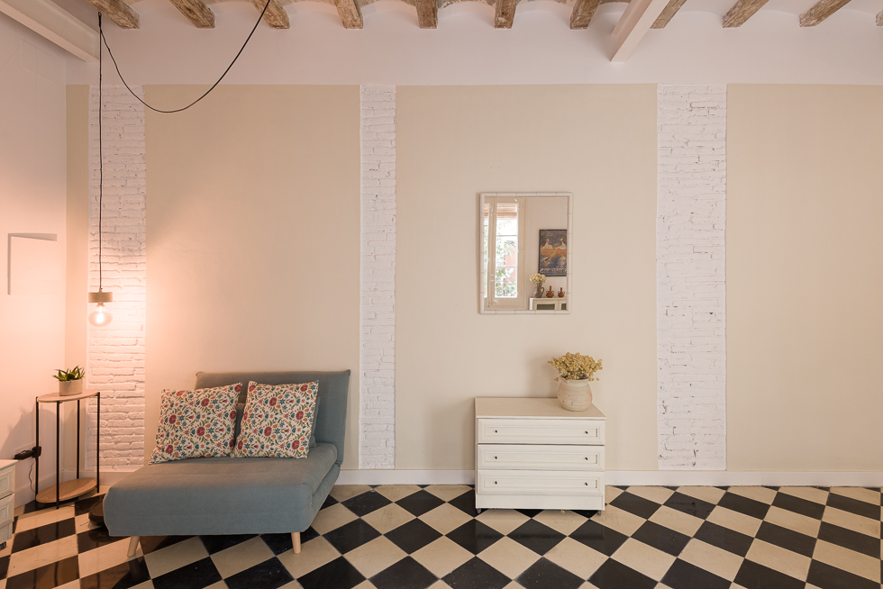 Cozy corner with a blue armchair, floral pillows, a small table, a white dresser, a square mirror on the wall reflecting the door onto the patio and checkered floor tiles.