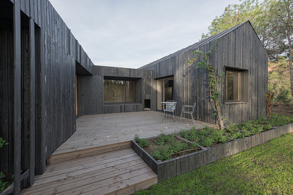 Outdoor view of a modern house with a pitched roof, dark wood cladding, a wooden deck, and a small garden area with greenery.