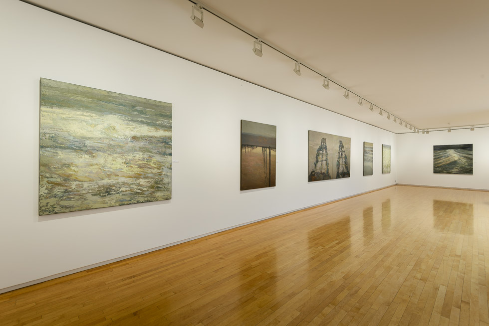 Spacious gallery room with wooden floors displaying large contemporary paintings on white walls, illuminated by track lighting.