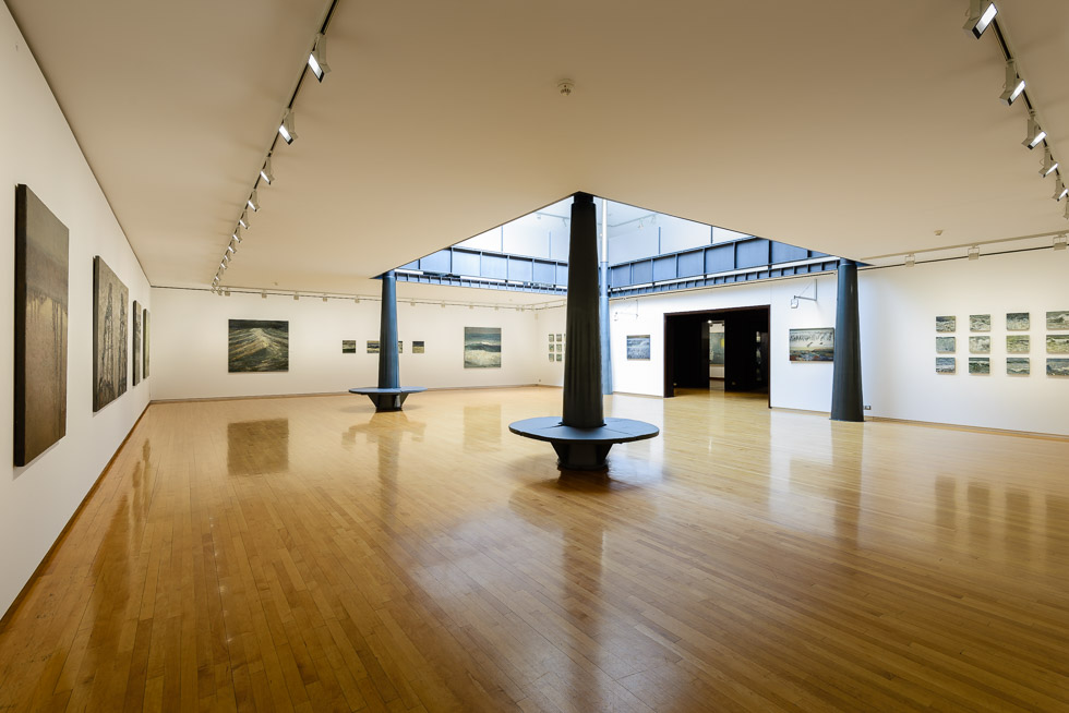 Art gallery with polished wooden floors, featuring large seascape paintings on white walls, centered around a skylighted atrium.