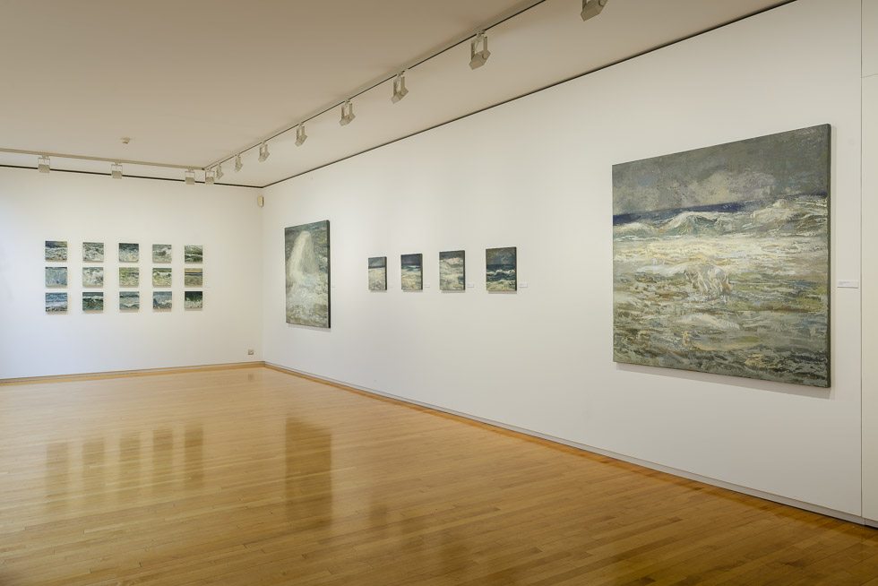 Art gallery with white walls and polished wooden floors, featuring an exhibition of seascape paintings. The artworks vary in size and are hung in a linear arrangement, illuminated by track lighting on the ceiling.