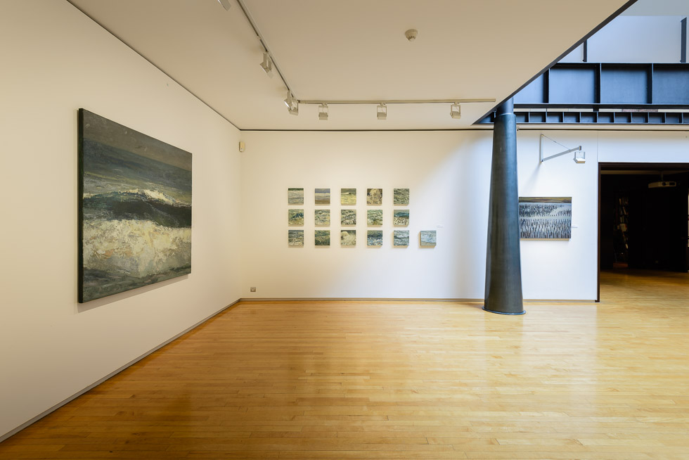 A spacious art gallery with white walls and polished wooden floors, displaying seascape paintings under track lighting. A black conic steel column stands in the side.