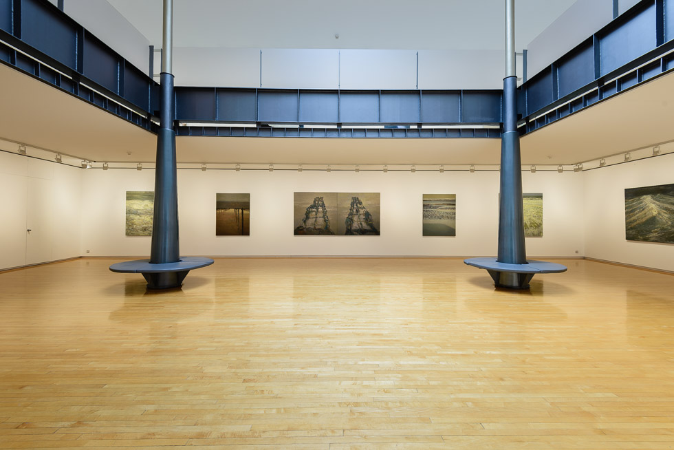 A spacious art gallery with polished wooden floors, white walls, and seascape paintings. Two black columns with circular benches stand in the side of a skylight-covered atrium.