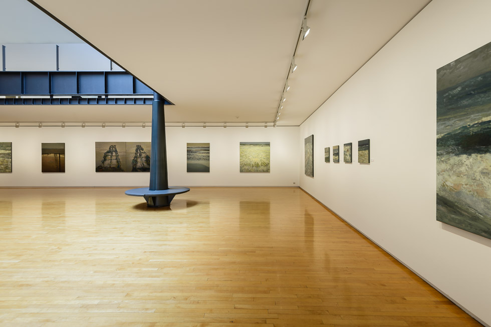 Art gallery with wooden floors, white walls, seascape paintings, and a black column with a circular bench in the left side.