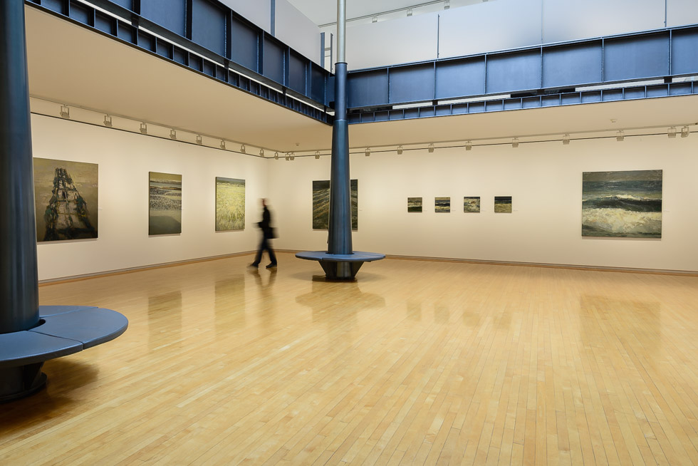 Art gallery with wooden floors, white walls, seascape paintings, and two black columns with a circular bench, featuring a blurred person walking on the background.