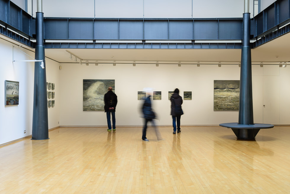 Art gallery with wooden floors, white walls, seascape paintings, and black columns, featuring people viewing the artwork.