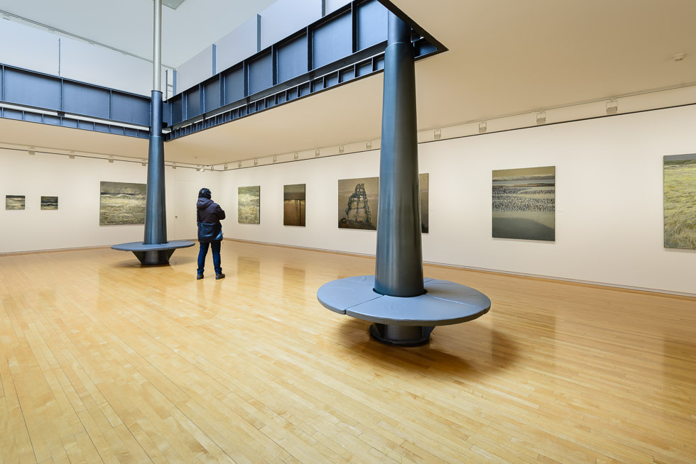 Art gallery with wooden floors, white walls, seascape paintings, and black columns, featuring a person viewing the artwork.