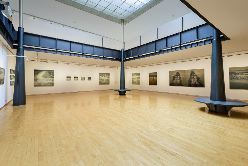 Spacious art gallery with a skylight atrium, displaying various seascape paintings on white walls, featuring wooden floors and metal columns.