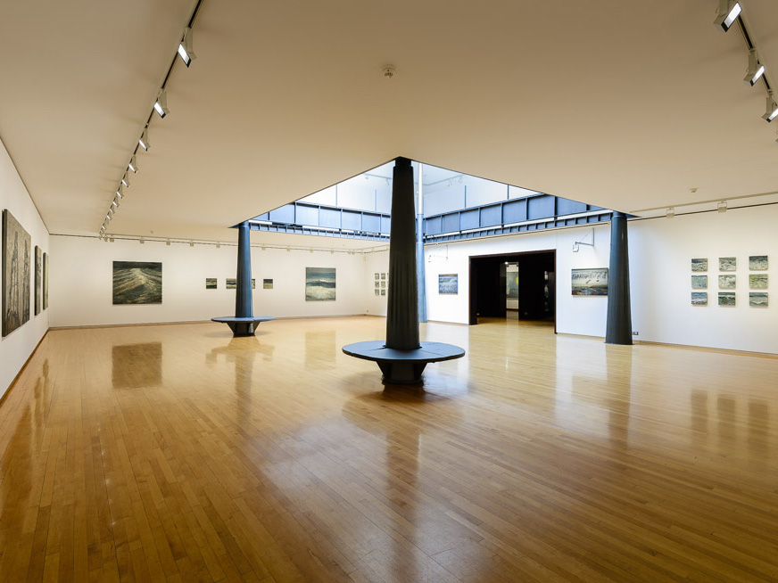 Art gallery with polished wooden floors, featuring large seascape paintings on white walls, centered around a skylighted atrium.