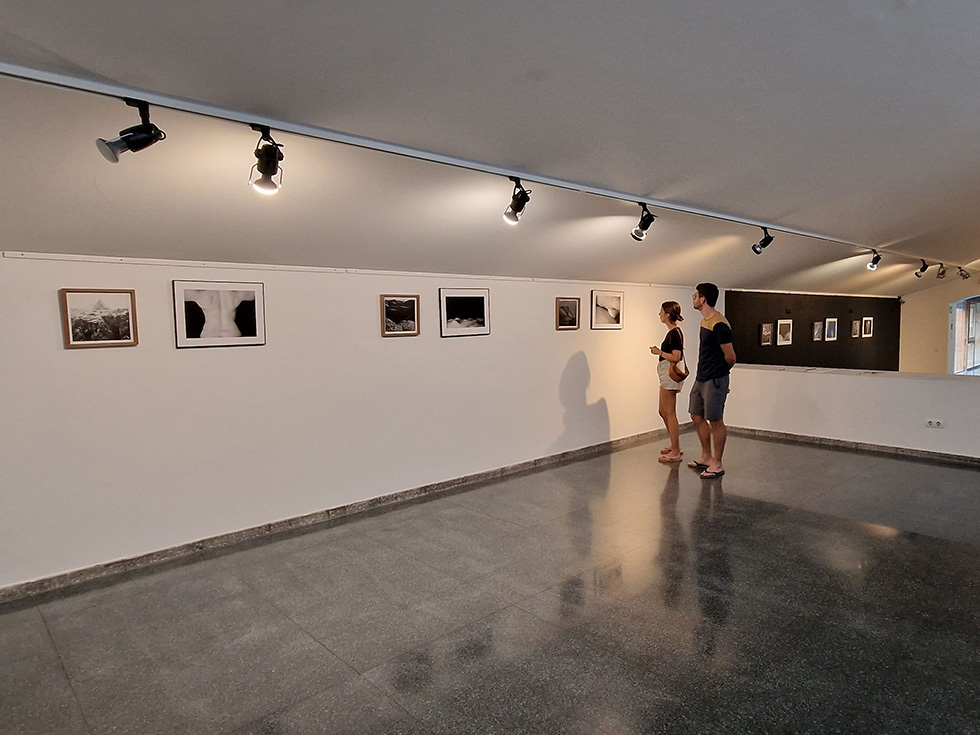 Two people viewing framed photographs on a gallery wall under spotlights.