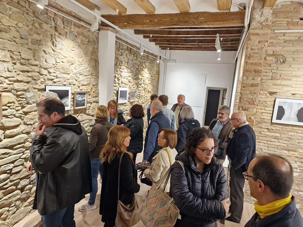 People attending an art exhibition opening at the Tres-e-u photography gallery, viewing photographs displayed on a rustic stone wall, and talking to each other.