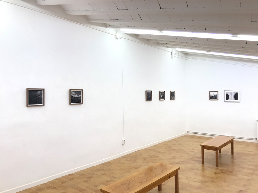 A minimalist gallery space with black-and-white photographs displayed on a white wall and wooden benches for seating.