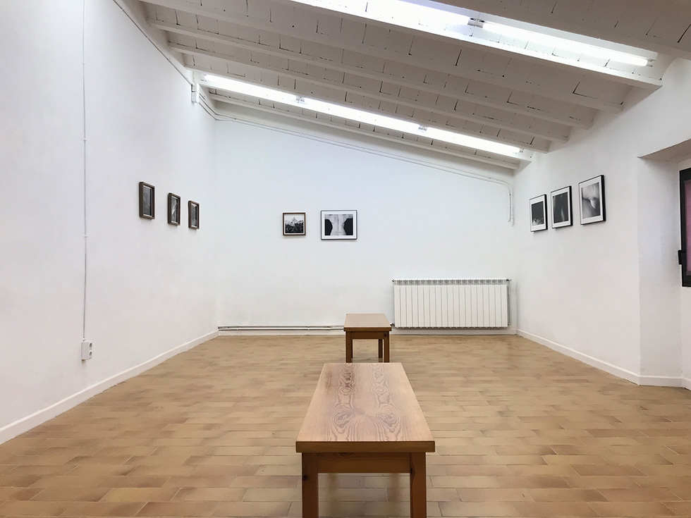 A minimalist gallery room with black-and-white photographs on white walls and two wooden benches in the center.