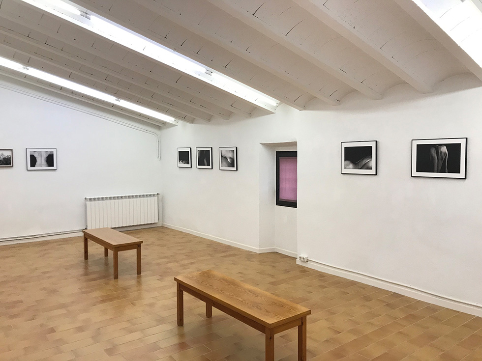 A minimalist gallery room with black-and-white photographs on white walls, two wooden benches in the center and a small window on one wall.