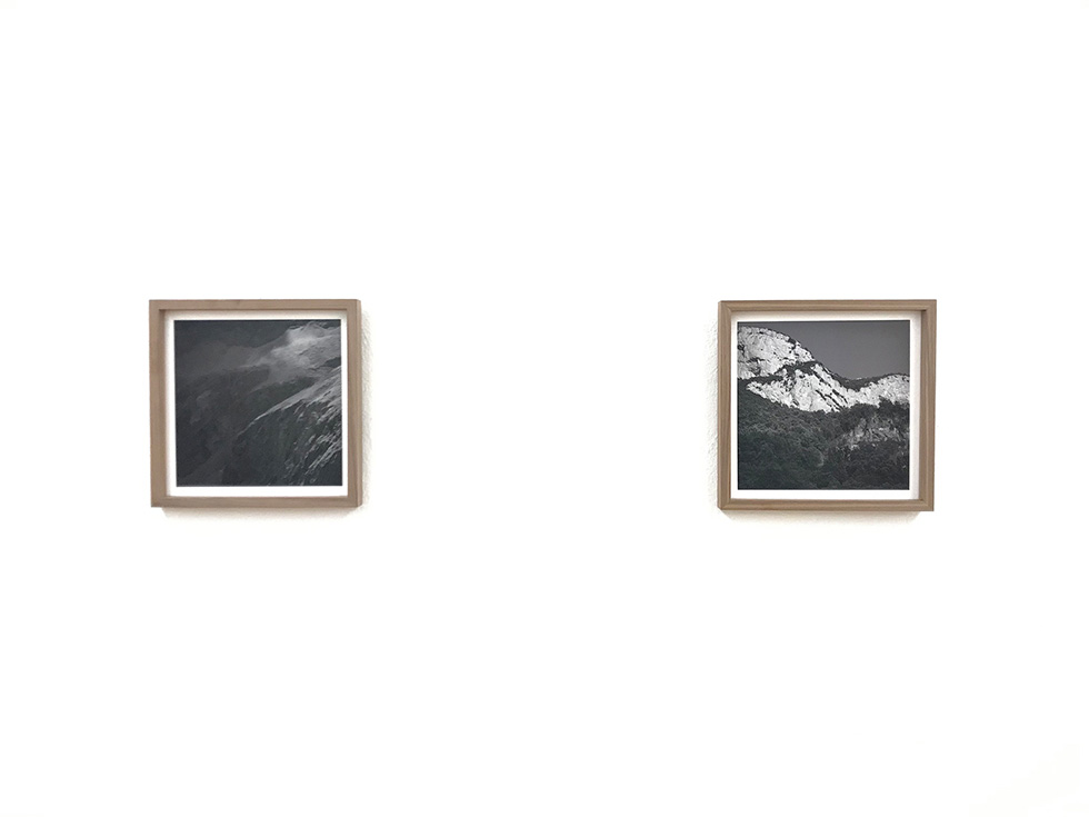 Two framed black-and-white photographs of mountains displayed on a white wall in a minimalist gallery setting.