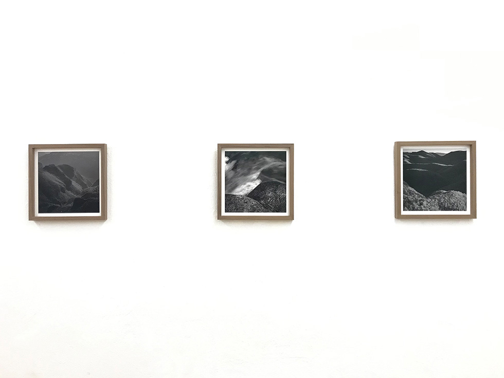 Three framed black-and-white photographs of mountain landscapes displayed on a white wall in a minimalist gallery setting.