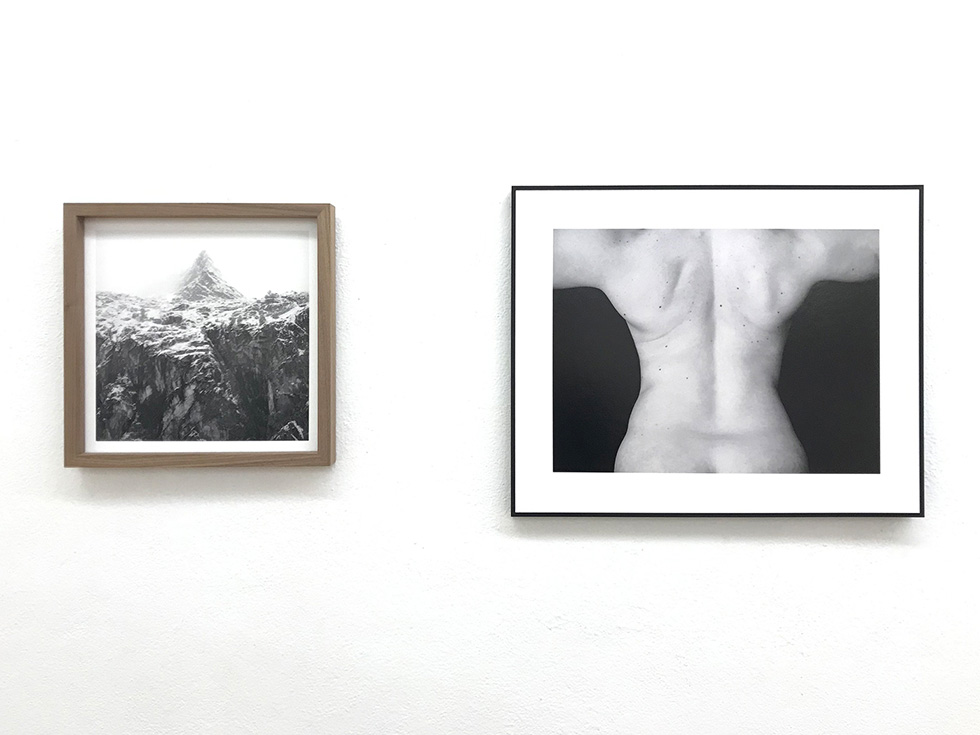 Two framed black-and-white photographs: one of a mountain peak and one of a person's back, displayed on a white gallery wall.