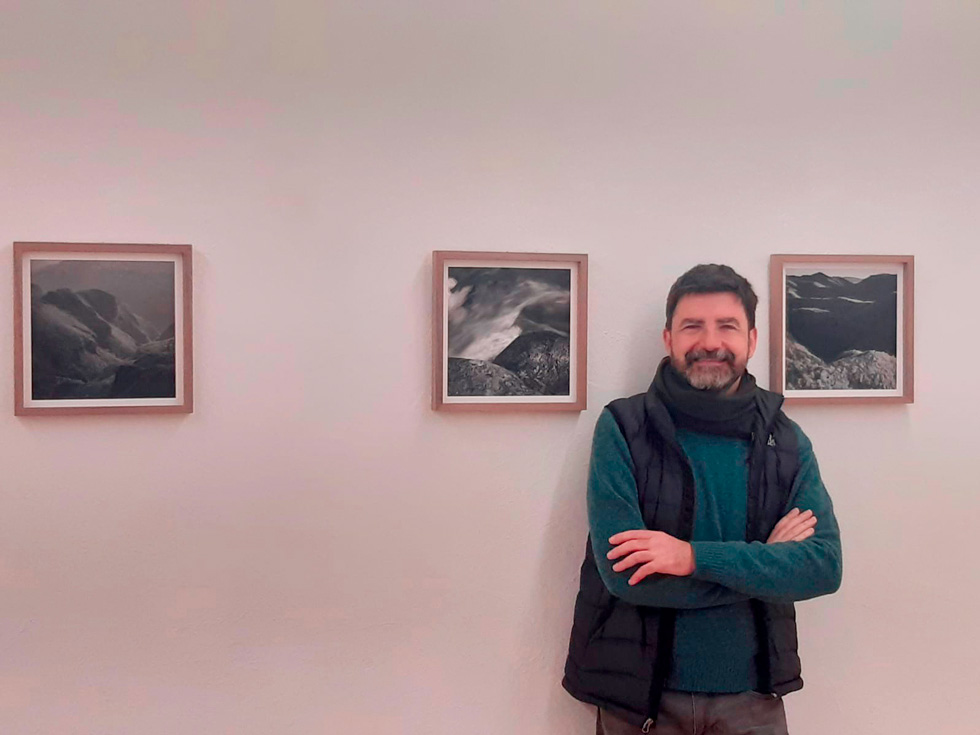 A man standing in front of three framed photographs of landscapes, smiling with arms crossed.