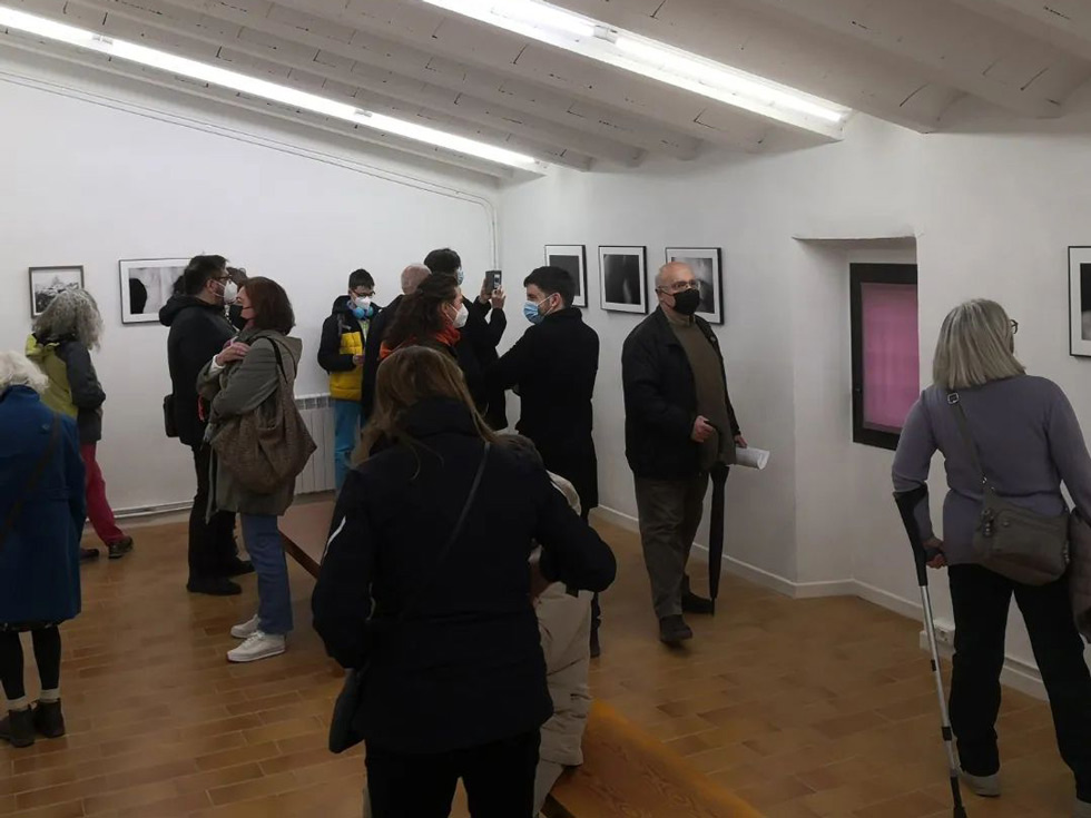 Visitors viewing framed photographs on the walls of an art gallery, all wearing surgical masks and engaging in conversation.