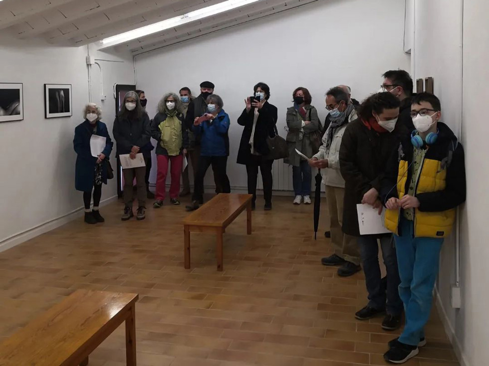 Visitors in an art gallery wearing surgical masks, some holding papers and taking photos, observing framed photographs on the walls.