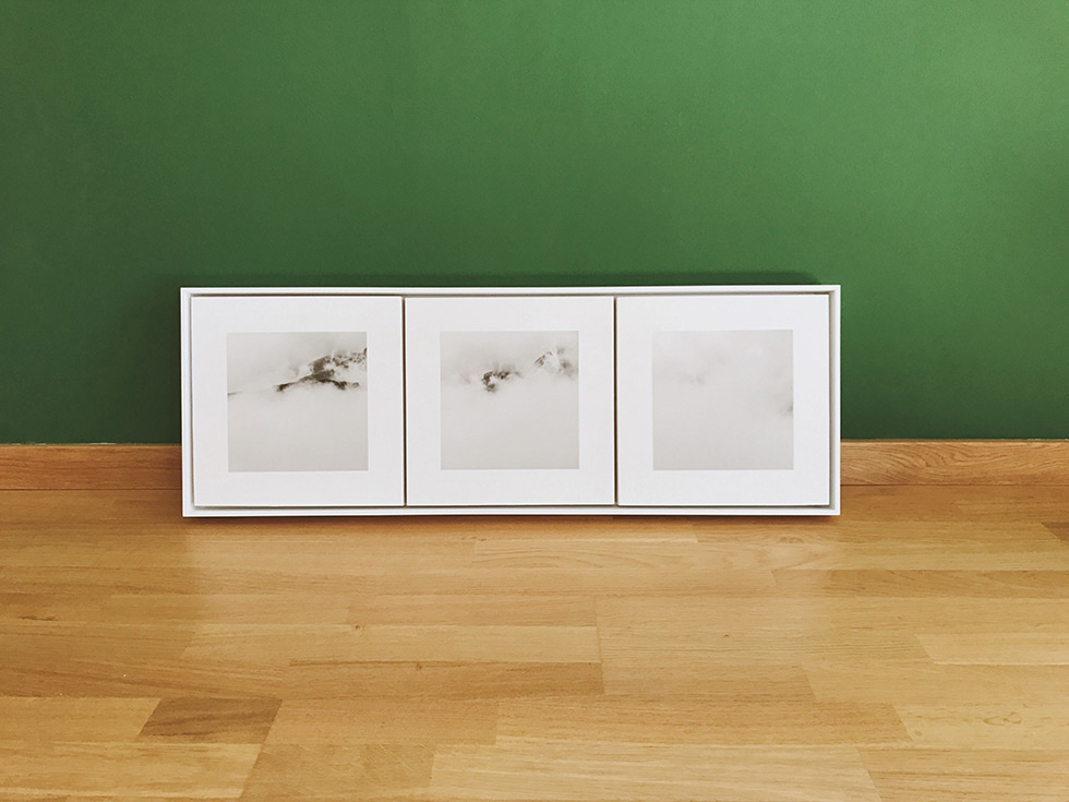 Framed triptych artwork featuring mountainous landscapes covered in fog displayed against a green wall on a wooden floor.