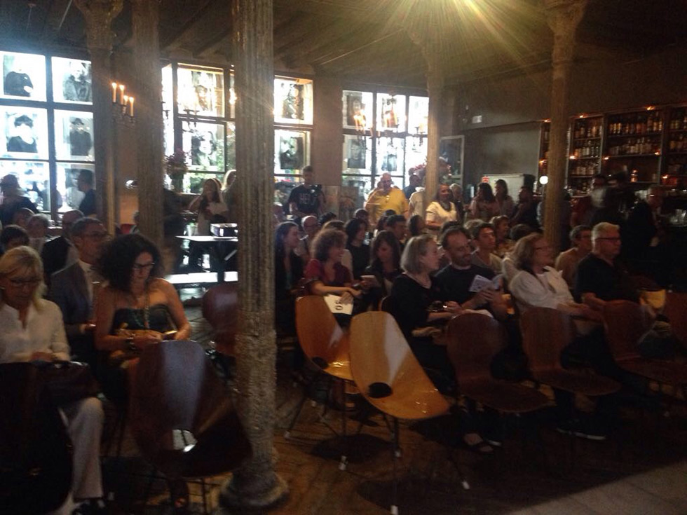 People seated in a dimly lit room, attending an event with a warm ambiance, surrounded by artistic decor and vintage elements.