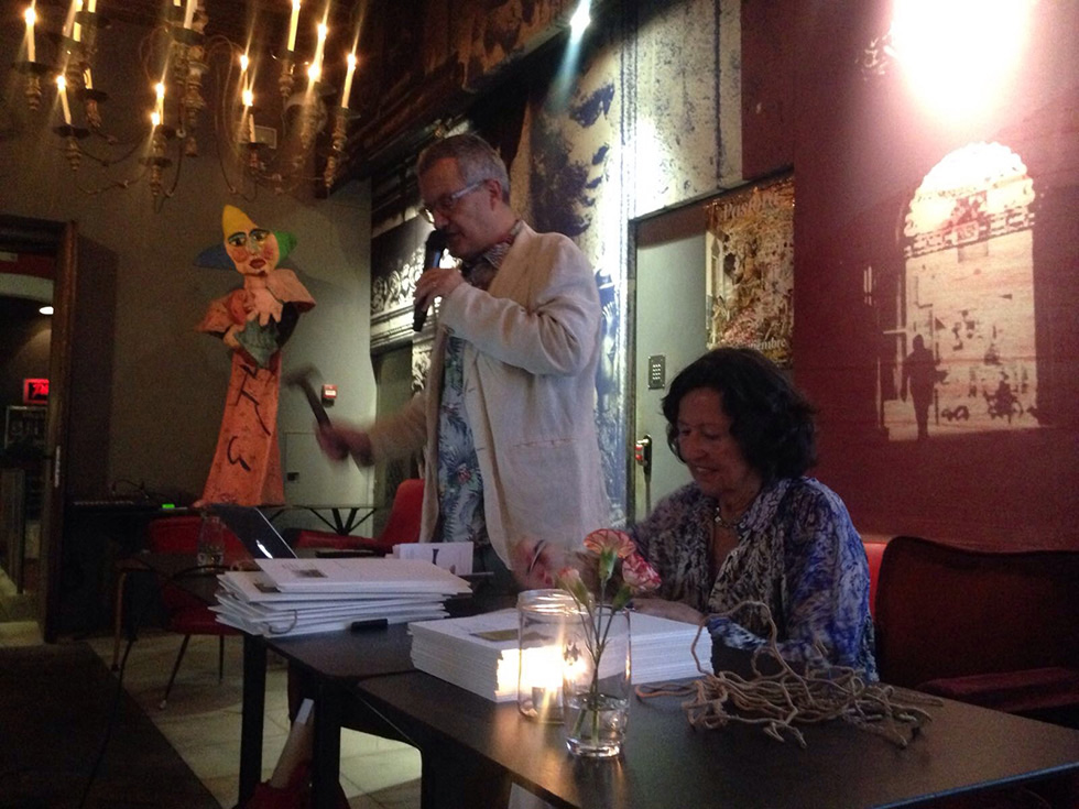 A man speaking into a microphone next to a seated woman at a table, with artistic decor and warm lighting in the background.