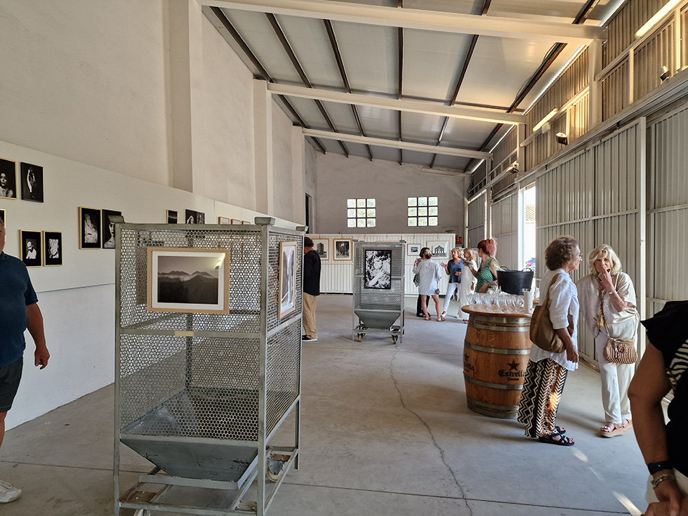 Interior of an exhibition space with high ceilings and exposed beams. Walls display framed photographs. Metal display stands and a wooden barrel are visible. Several visitors are standing and conversing in small groups throughout the space, giving the impression of a gallery opening.