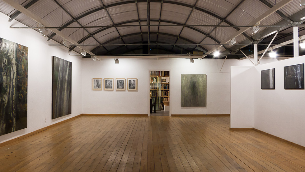 Exhibition space with wooden flooring, large artworks on walls, and a doorway leading to a room with bookshelves.