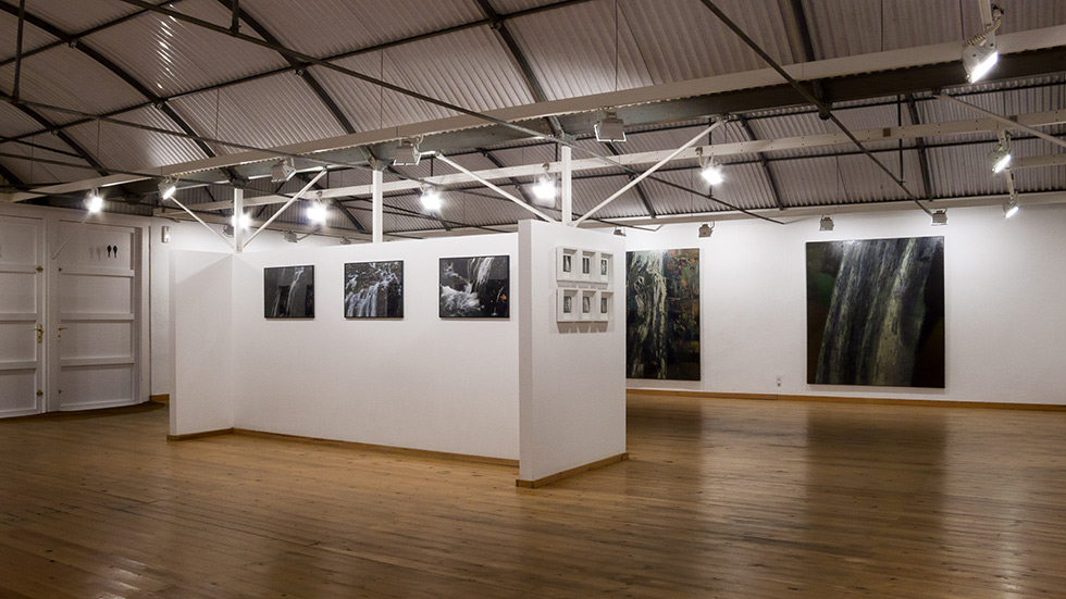 Spacious gallery with artworks on walls, wooden floors, and exposed metal ceiling.