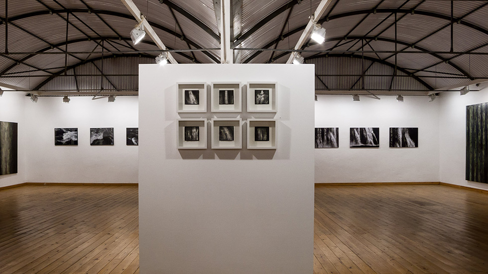 Gallery interior with framed artworks on walls, centered display of smaller frames, wooden floors, and vaulted metal ceiling.