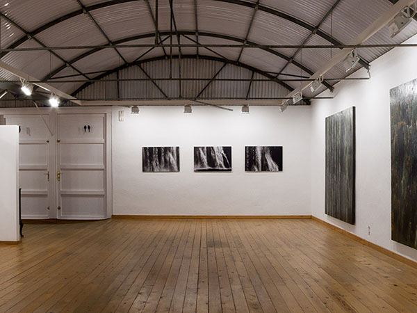 Spacious gallery with artworks on walls, wooden floors, and exposed metal ceiling.