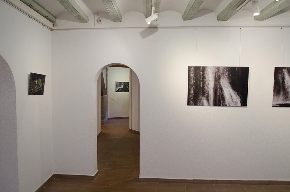 Archway view of gallery with framed photographs on white walls, wooden ceiling beams, and tiled floor.