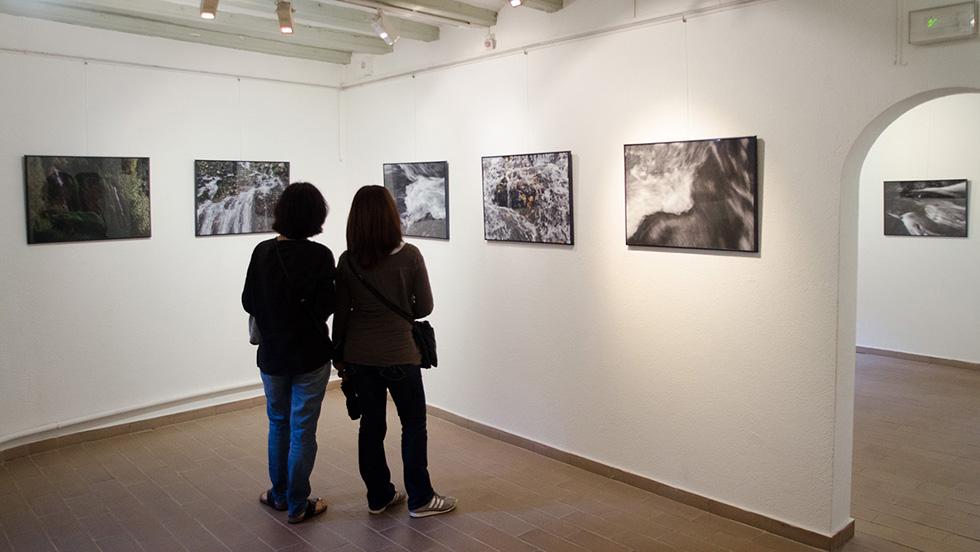 Two people viewing a series of waterfall photographs on a white gallery wall, with wooden ceiling beams and tiled floor.