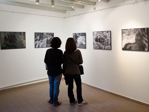 Two people viewing a series of waterfall photographs on a white gallery wall, with wooden ceiling beams and tiled floor.