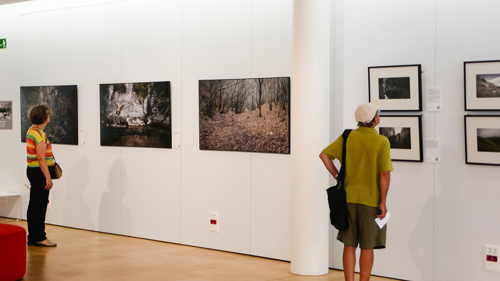 Two people viewing photographs in an art gallery, with large unframed images on the left wall and smaller framed photos on the right.