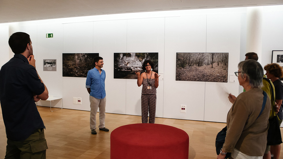 A group of people listening to a speaker in an art gallery, with large landscape photographs displayed on the wall behind them.