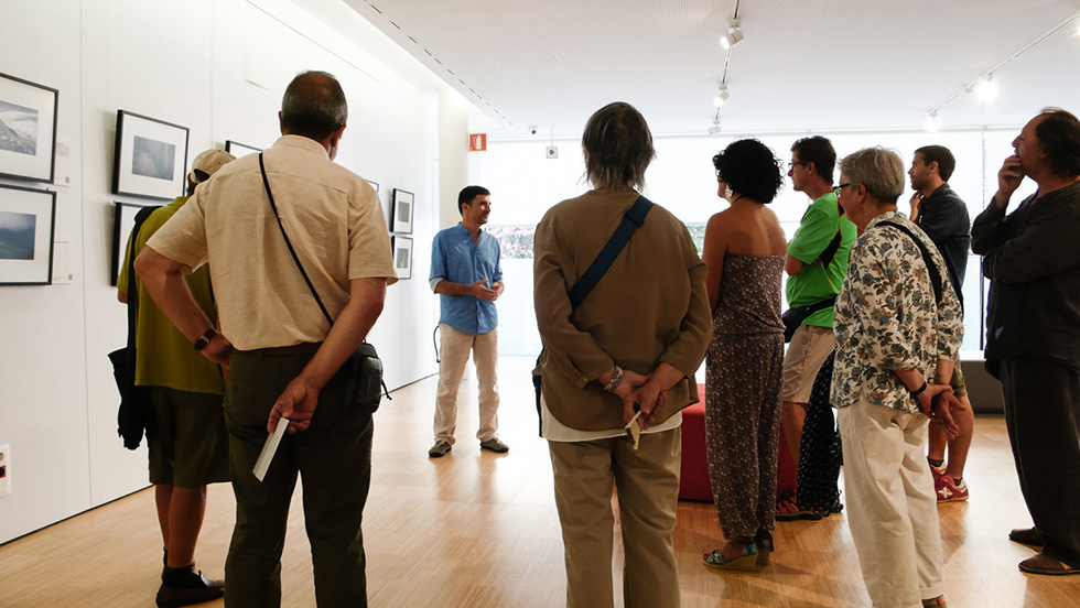 A group of people attentively listening to a speaker in an art gallery, with framed photographs displayed on the wall behind them.