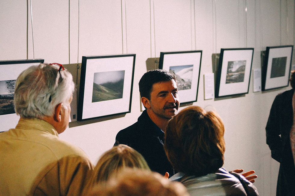 A man speaking to a group of people in front of a wall displaying framed photographs.