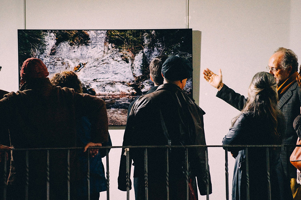 People standing in front of a large color photograph at an exhibition, one man gesturing towards the photo.