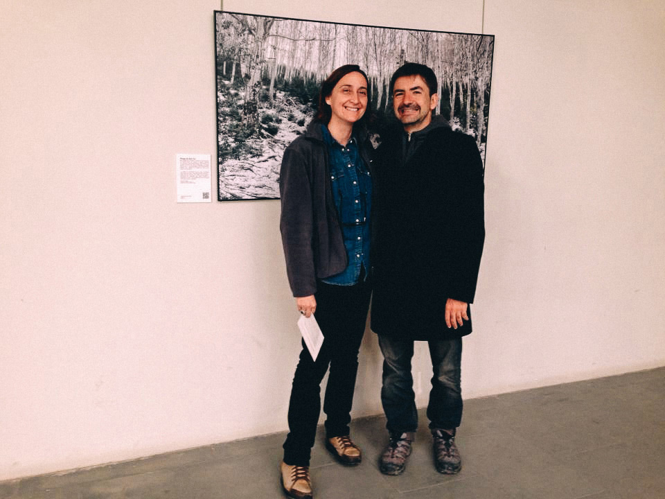 Two people standing in front of a large photograph of a snowed forest, smiling at the camera.