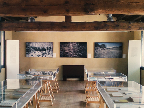 View into a gallery room with wooden beams, framed photos on the walls and display cases on wooden stands.