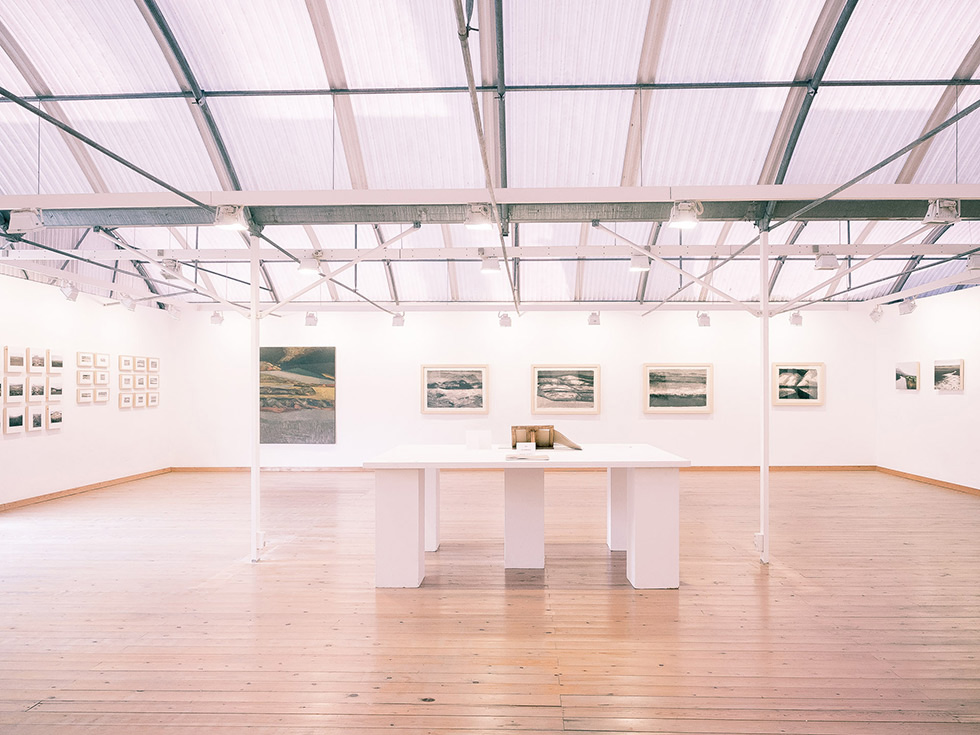 Bright exhibition hall with framed artworks on the walls, a high curved ceiling, wooden floors, and a central white table displaying books.