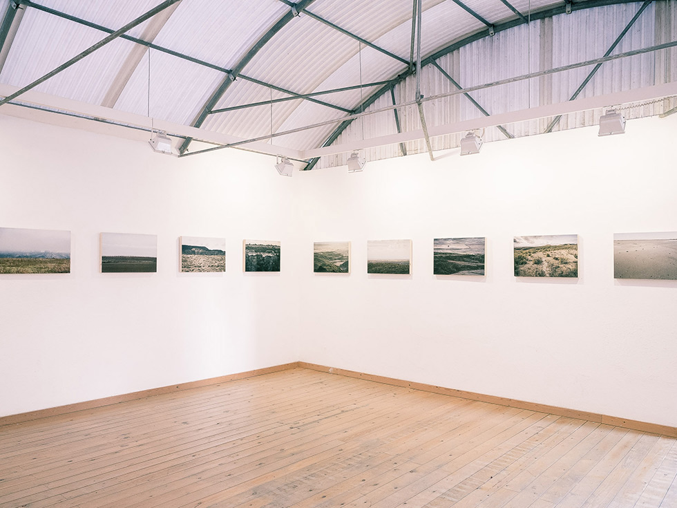 Exhibition hall with a series of landscape photographs arranged in a single row along the white walls, featuring a high curved ceiling and wooden floors.