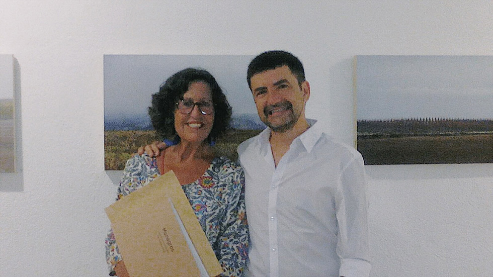 Two people standing in front of a wall with landscape photographs, smiling at the camera, one holding a book. The background shows a calm, serene landscape in the photographs.