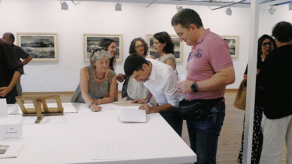 People gathered in an art gallery, engaging with the art and each other. One person is signing a book on a table, while others are discussing and viewing landscape photographs displayed on the walls in the background. The atmosphere is lively and interactive.