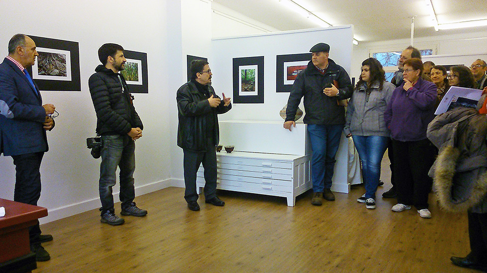 A group of people gathered indoors, listening to a person speak at a photo exhibition.